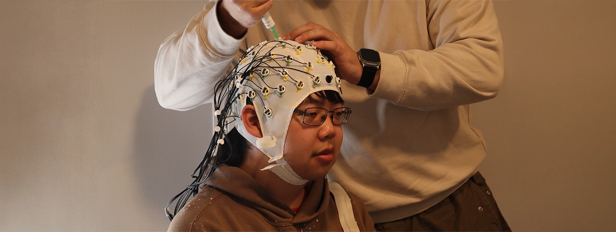 The image shows a person wearing a white EEG (electroencephalogram) cap with multiple electrodes, while another person applies conductive gel or paste to the scalp through the cap's openings using a blunt-tip syringe or dispenser.