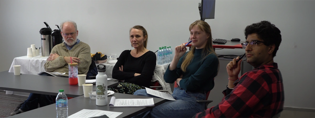 The image shows four people sitting at tables in a meeting or workshop room.