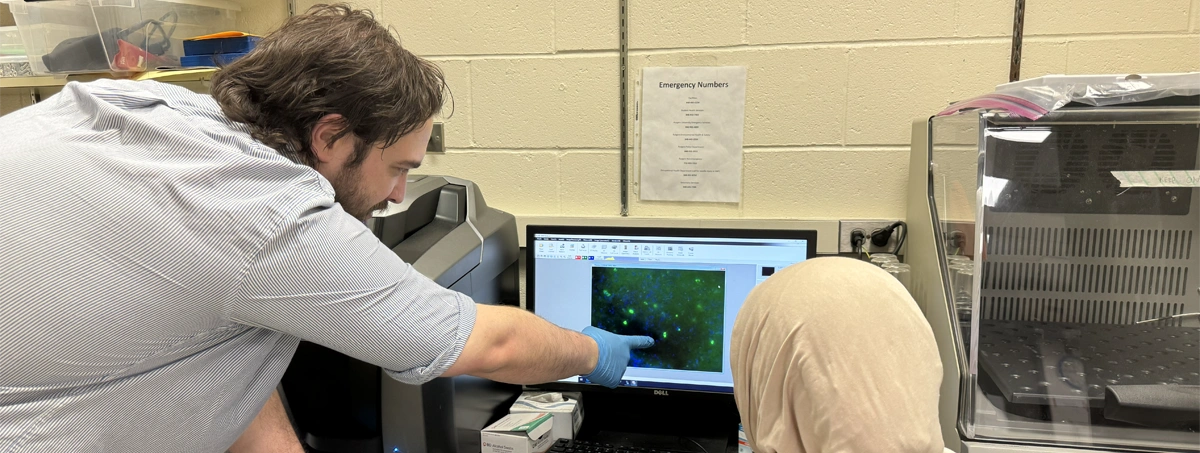 The image shows two people in a laboratory looking at a computer screen displaying fluorescence microscopy data of cells.
