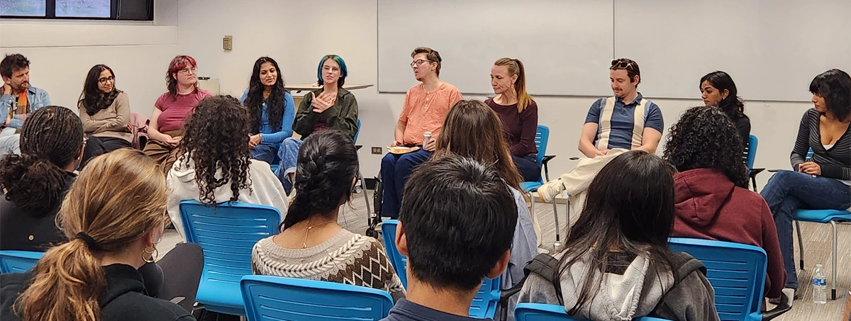 The image shows a group of people sitting in a circle in a classroom or meeting room, engaged in a discussion or panel session.