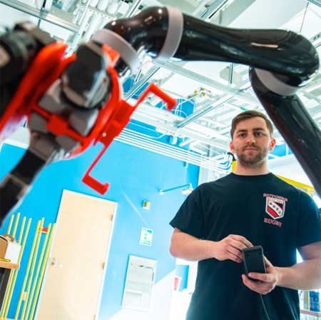 This image shows a person wearing a black t-shirt with a Rutgers logo, holding a controller near a robotic arm.
